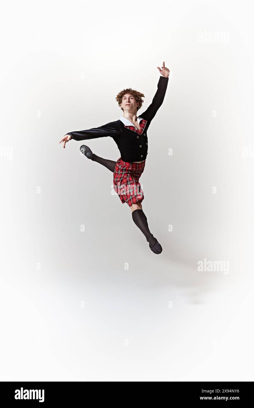 Jeune danseur de ballet masculin, vêtu d'un kilt de tartan traditionnel et d'une tenue noire, effectue un saut de ballet impressionnant sur fond blanc de studio. Banque D'Images