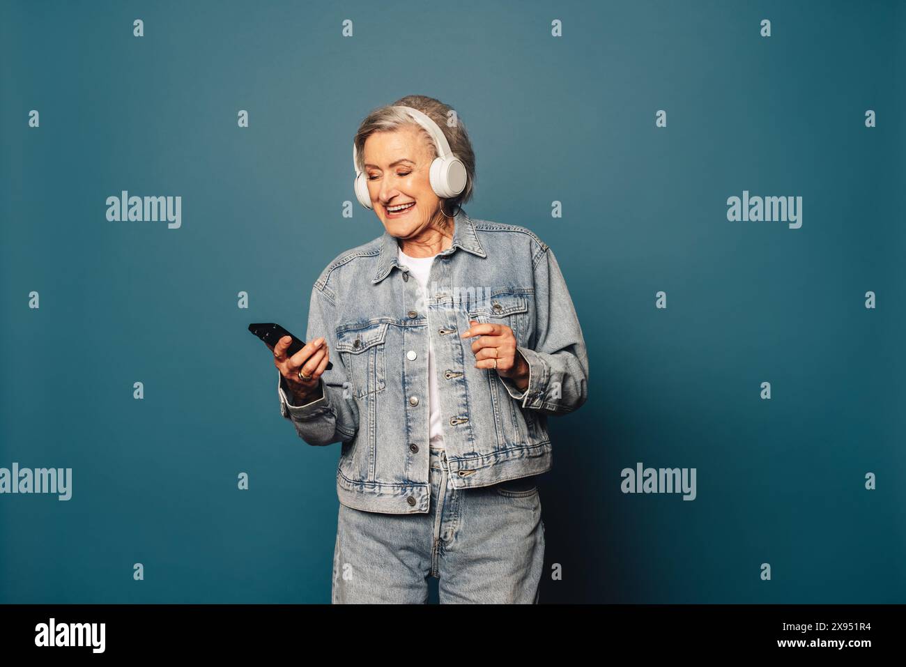 Femme joyeuse et à la mode aux cheveux gris danse et sourit dans un studio, portant une tenue en denim. Tenant un smartphone, elle écoute de la musique. Banque D'Images
