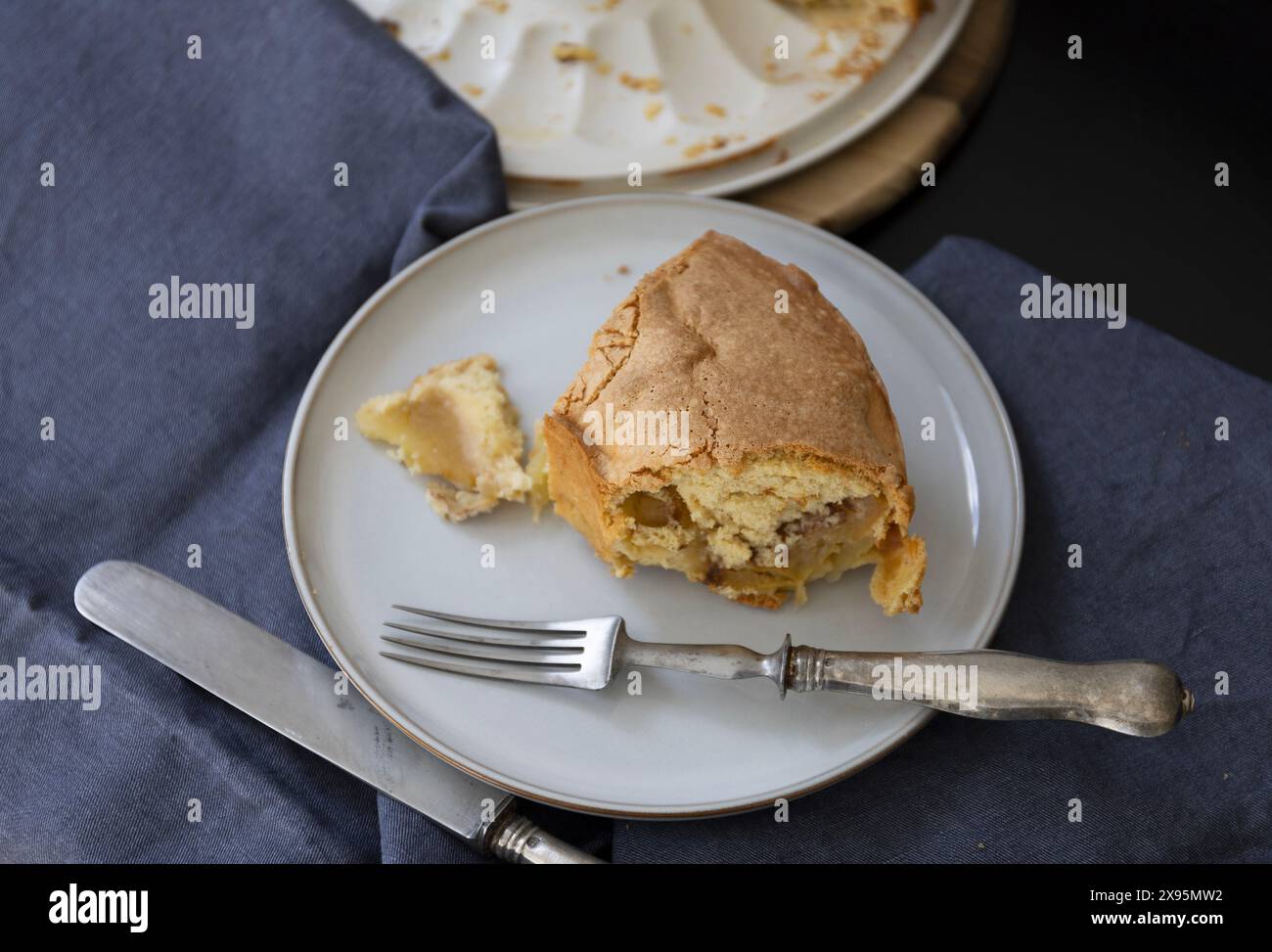 Un morceau de gâteau éponge sur une assiette de couleur claire avec fourchette vintage et couteau sur un fond noir avec draperie bleu foncé à la lumière naturelle Banque D'Images