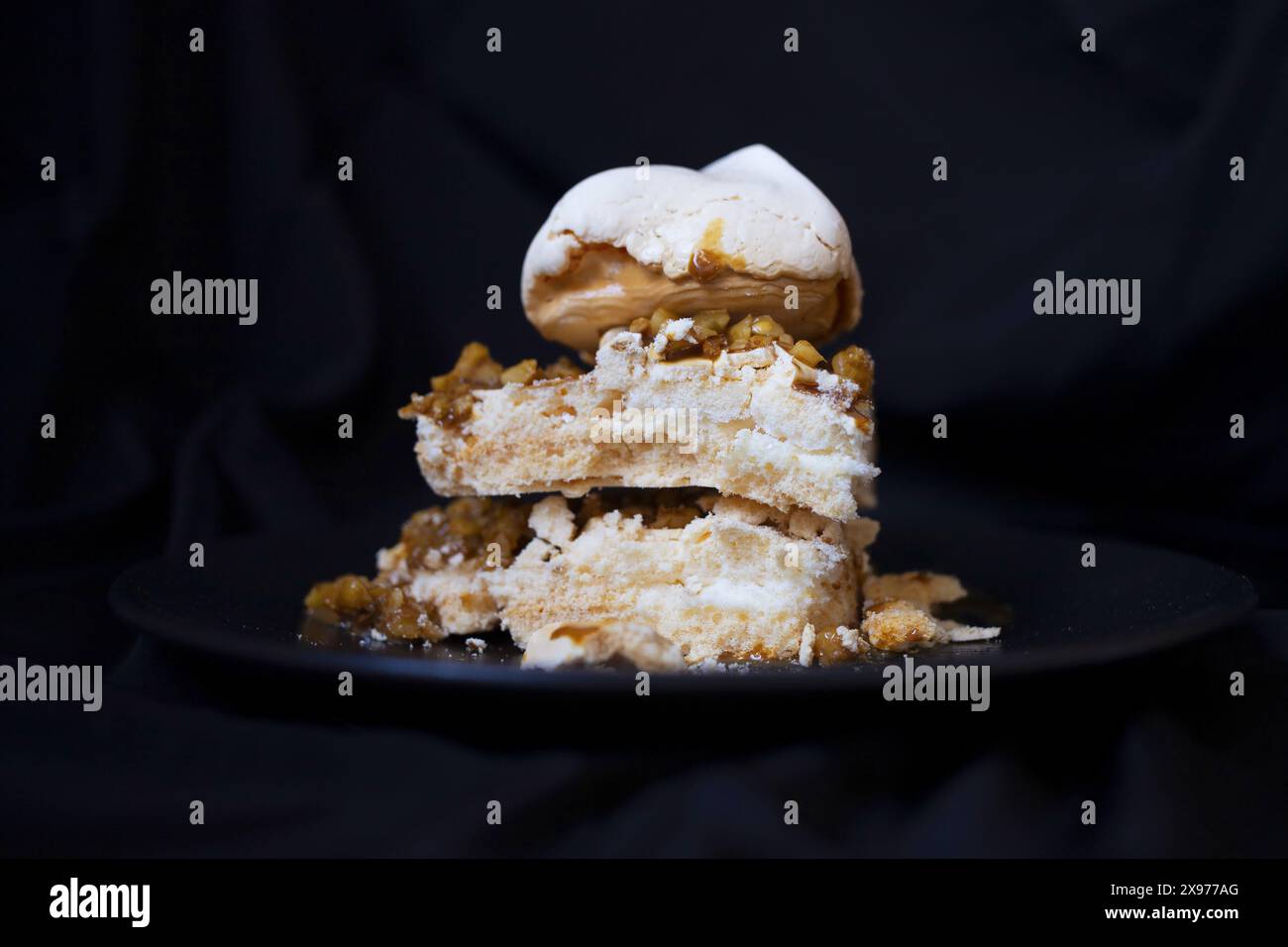 Photographie alimentaire basse clé d'un morceau de gâteau de meringue Pavlova avec sauce au café et noix sur une plaque de céramique noire sur un fond de tissu de soie sombre Banque D'Images