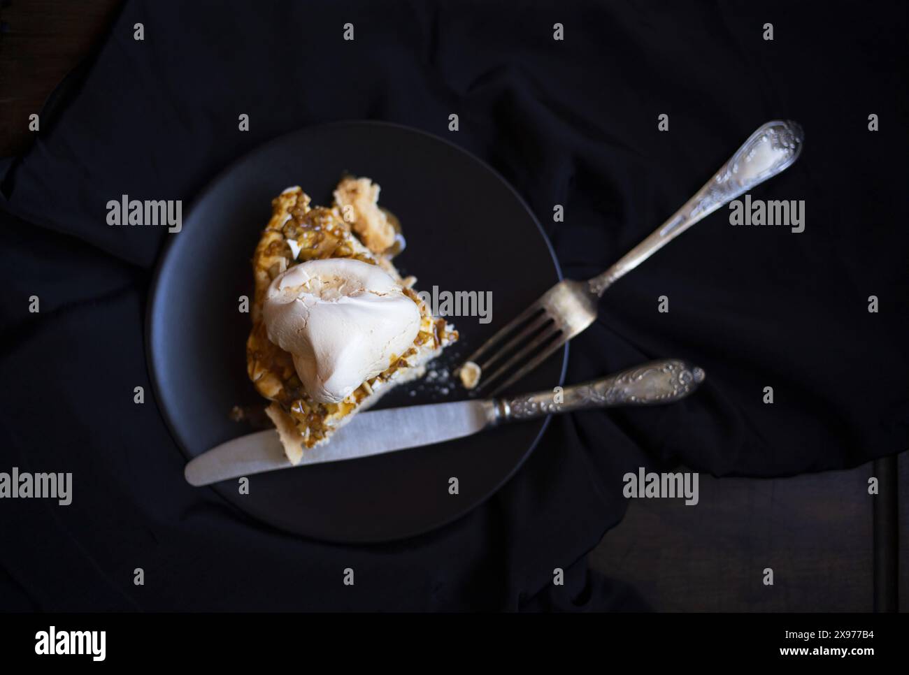 Photographie alimentaire basse clé de table d'un morceau de gâteau de meringue Pavlova avec sauce au café et noix sur une plaque de céramique noire avec fourchette vintage et kni Banque D'Images