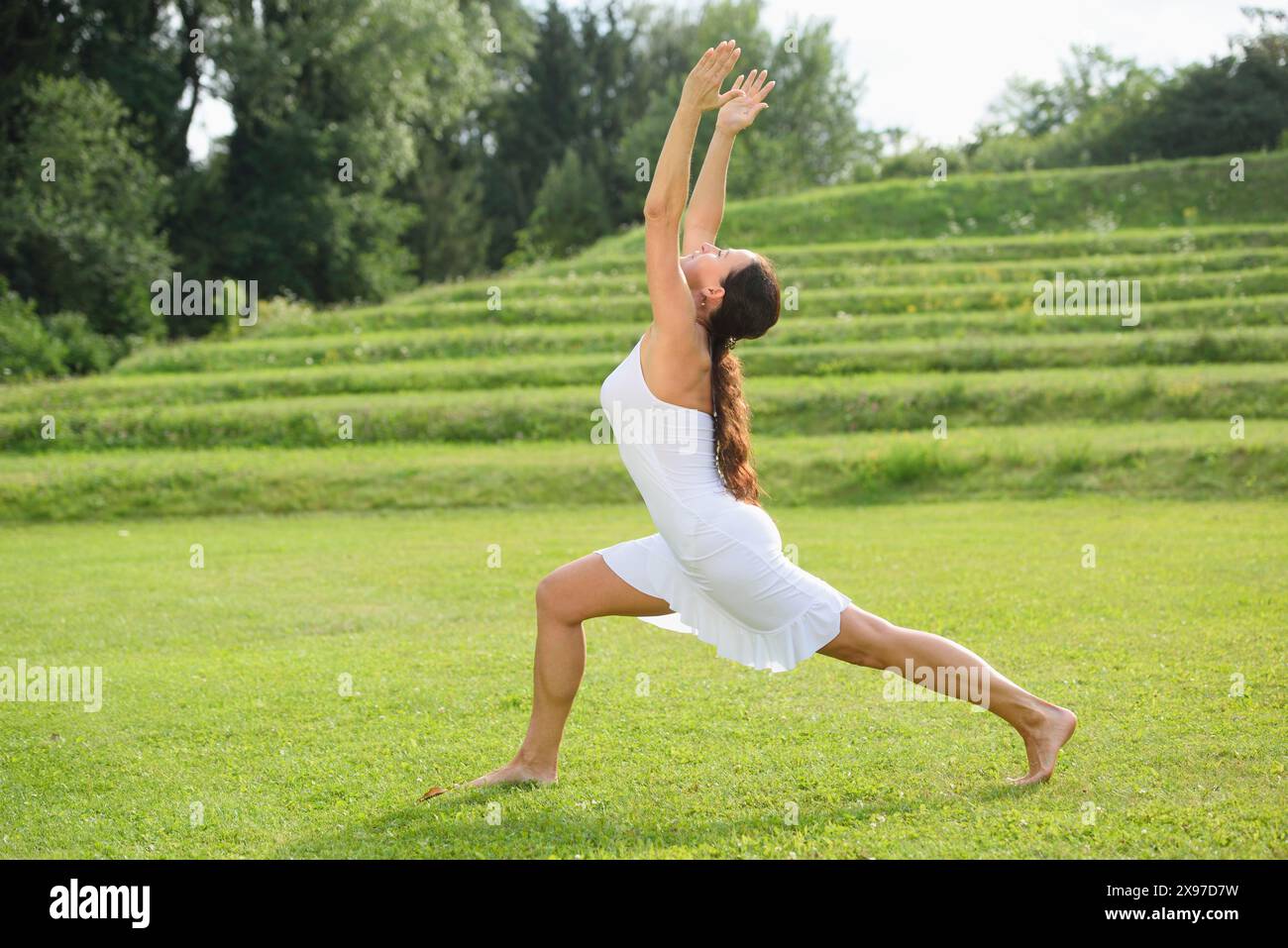 Femme d'âge moyen faisant du yoga, la posture du croissant ou la fente haute, Anjaneyasana, sur une prairie dans un parc en été, Allemagne Banque D'Images