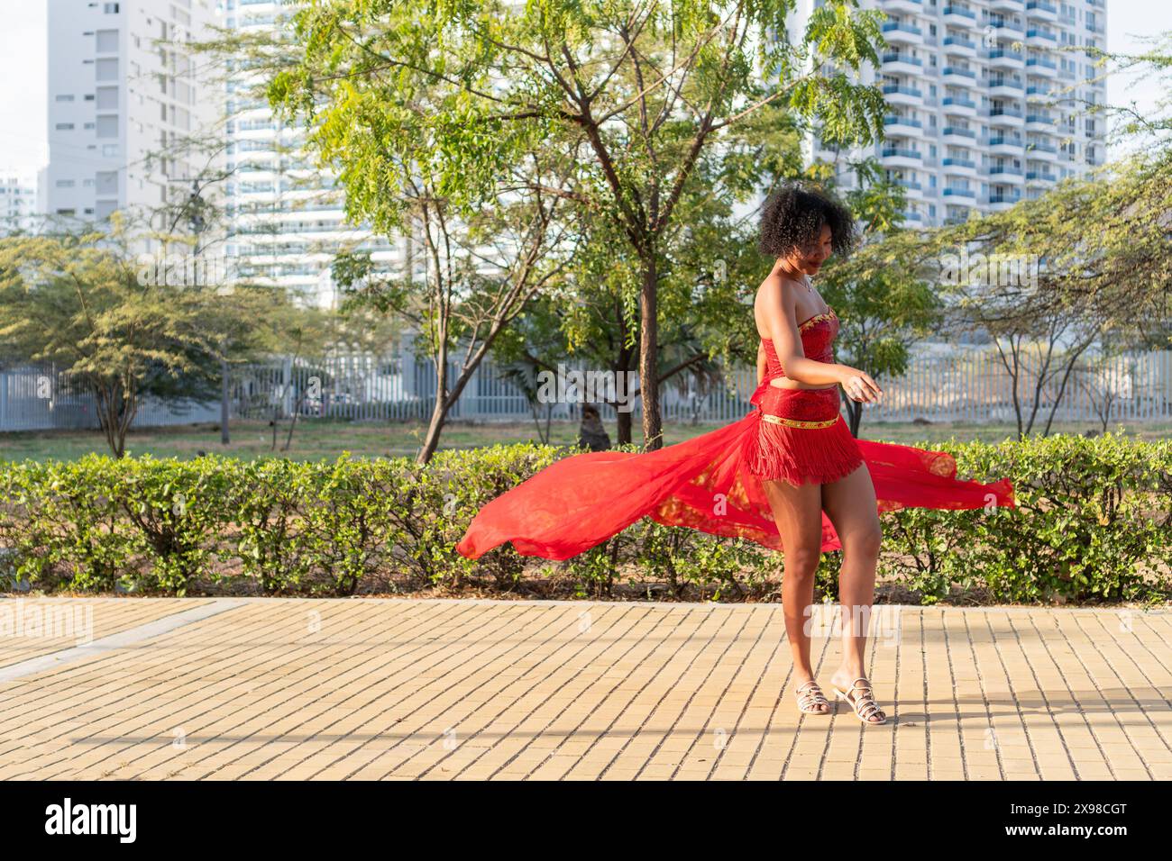 Une femme dans une robe rouge vibrante danse gracieusement dans un parc urbain avec de la verdure et des immeubles de grande hauteur en arrière-plan, mettant en valeur un moment animé. Banque D'Images
