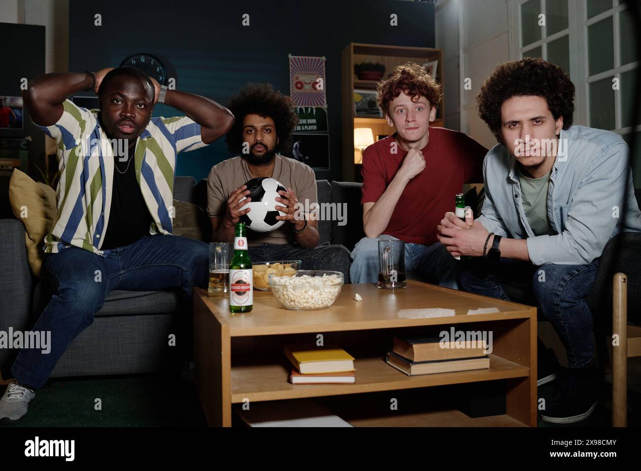 Groupe d'amis masculins ethniquement diversifiés assis dans le salon tard dans la soirée regardant un match de football à la télévision Banque D'Images