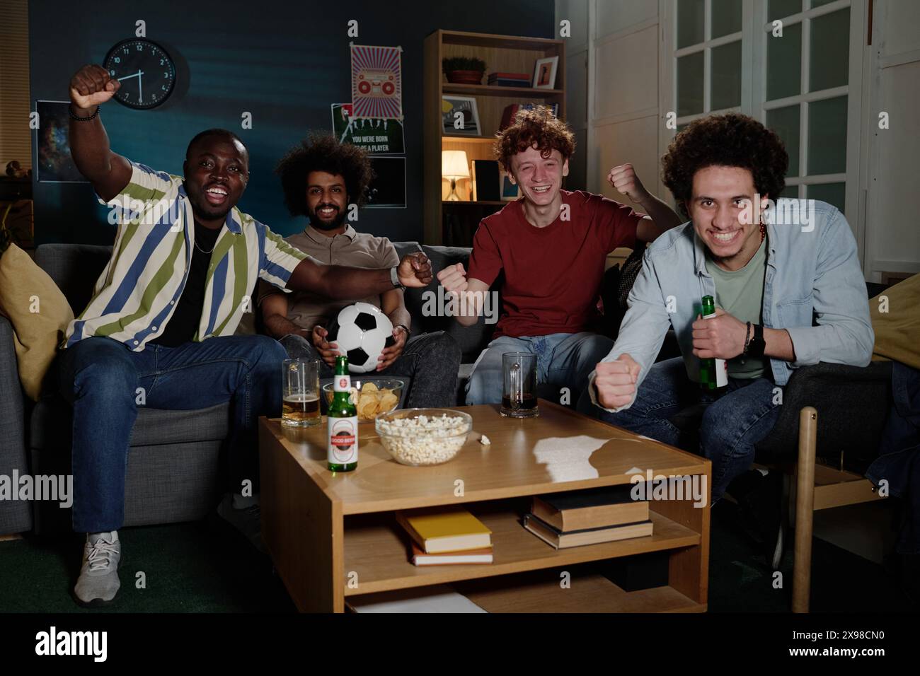 Groupe multi-ethnique de jeunes hommes joyeux assis dans le salon appréciant regarder un match de football à la télévision Banque D'Images