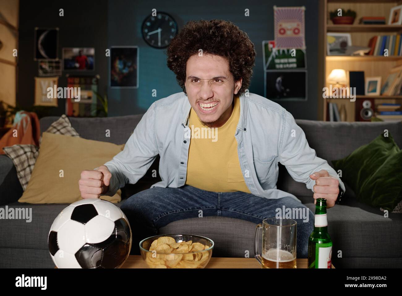 Plan moyen d'un jeune homme biracial excité assis sur le canapé dans le salon regardant un match de football à la télévision tard dans la soirée Banque D'Images
