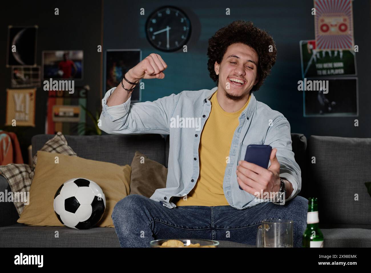 Plan moyen de joyeux gars biracial assis sur le canapé tard dans la soirée regardant un match de football en ligne sur smartphone, espace de copie Banque D'Images
