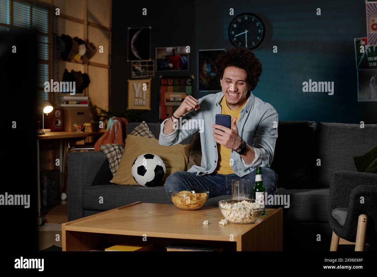 Joyeux jeune homme biracial assis sur le canapé dans le salon regardant le match de football en ligne sur smartphone, espace de copie Banque D'Images
