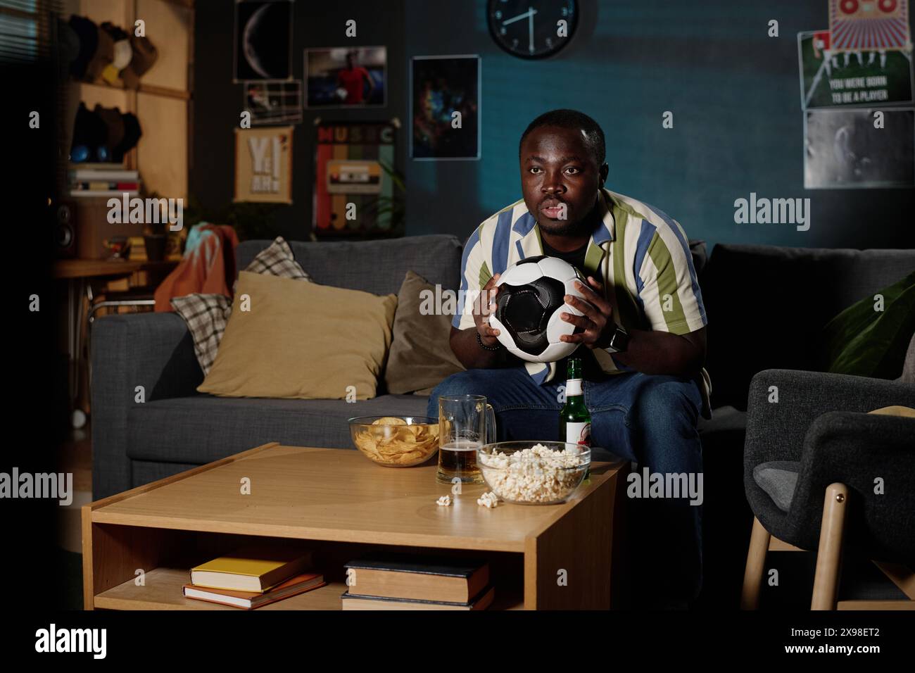 Jeune homme afro-américain tenant le ballon assis sur le canapé dans le salon tard dans la soirée regardant un match de football à la télévision Banque D'Images