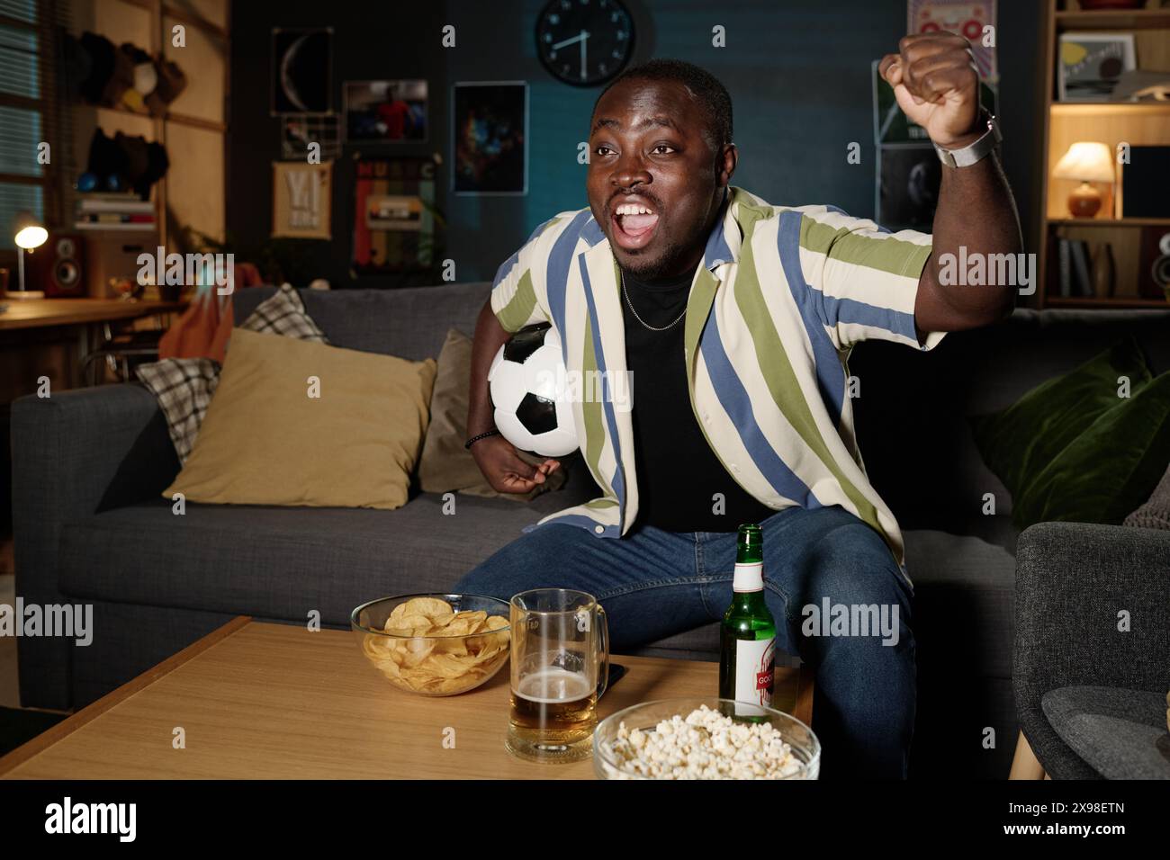 Plan moyen long de joyeux gars afro-américain tenant le ballon assis sur le canapé dans le salon regardant le match de championnat de football à la télévision Banque D'Images
