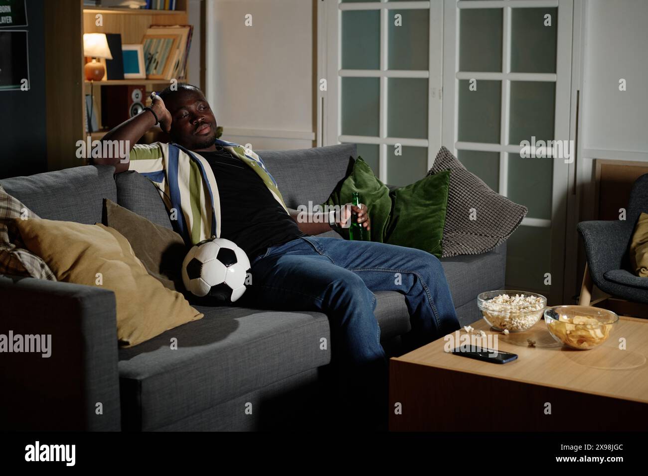 Jeune homme noir tenant une bouteille de bière assis détendu sur le canapé dans le salon regardant un match de football à la télévision, espace de copie Banque D'Images