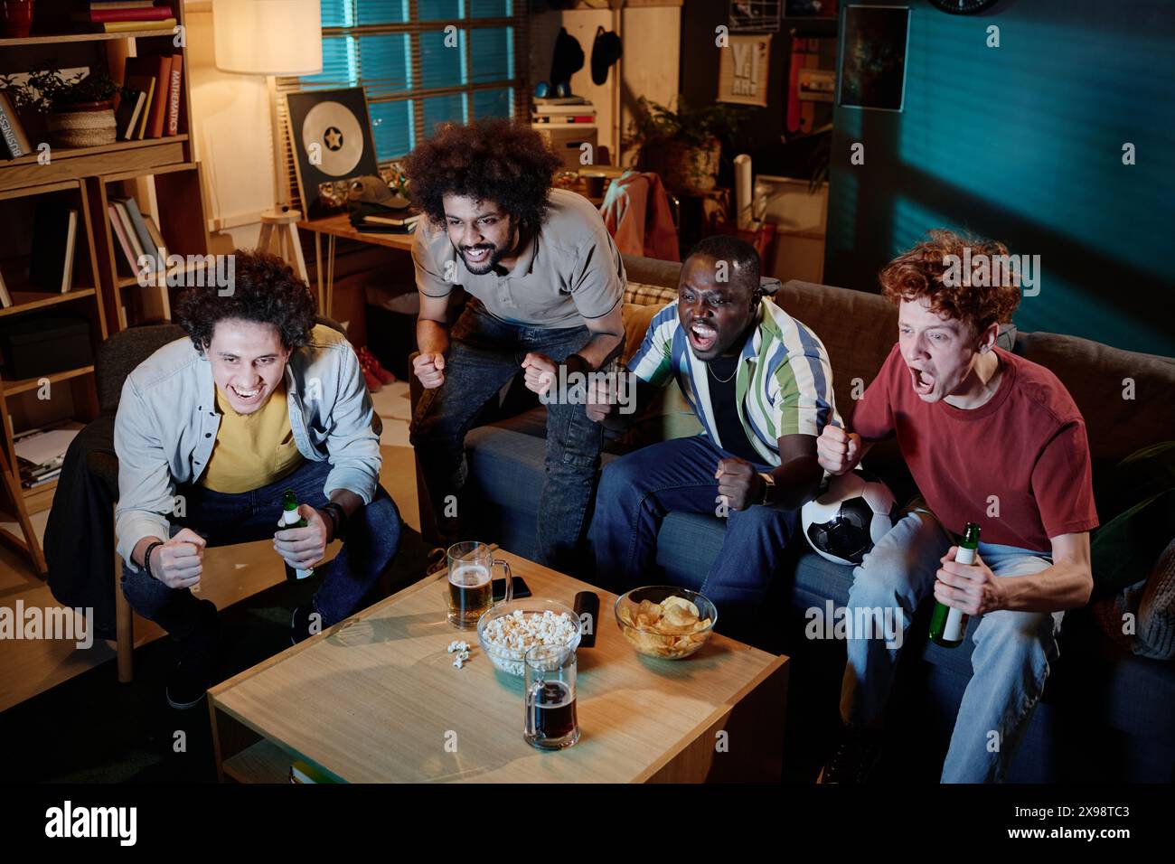 Vue en angle élevé d'un groupe multiethnique de quatre hommes appréciant de regarder un match de football à la télévision tard dans la soirée Banque D'Images