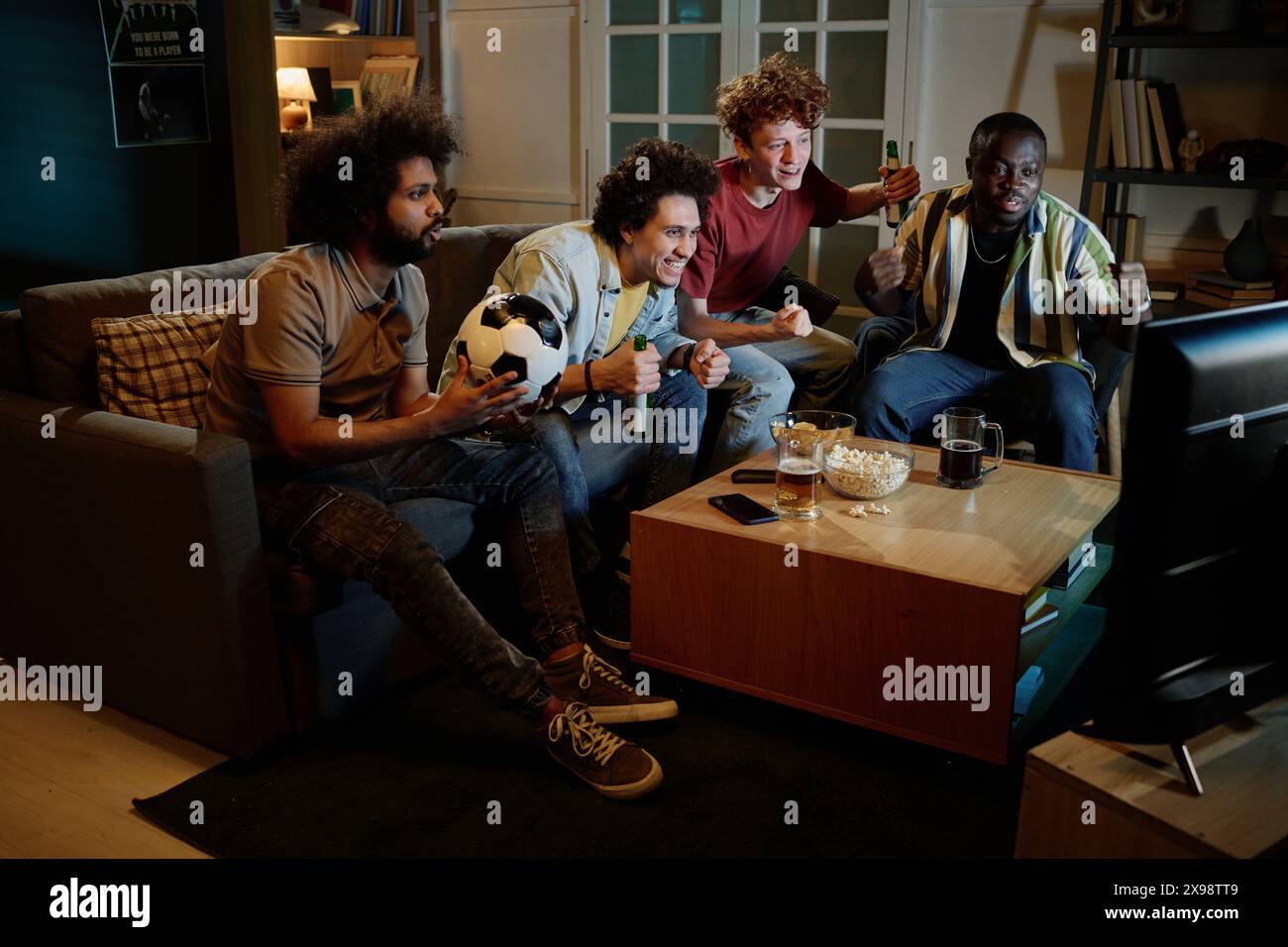 Groupe d'amis joyeux assis dans le salon s'amusant à regarder un match de football à la télévision tard dans la soirée Banque D'Images