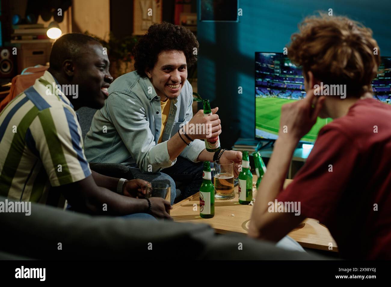 Jeunes hommes ethniquement diversifiés assis dans le salon discutant tout en regardant un match de football à la télévision tard dans la soirée Banque D'Images