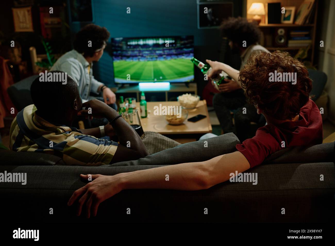 Vue arrière de quatre jeunes amis masculins passant la soirée à l'intérieur à regarder un match de football à la télévision et boire de la bière Banque D'Images