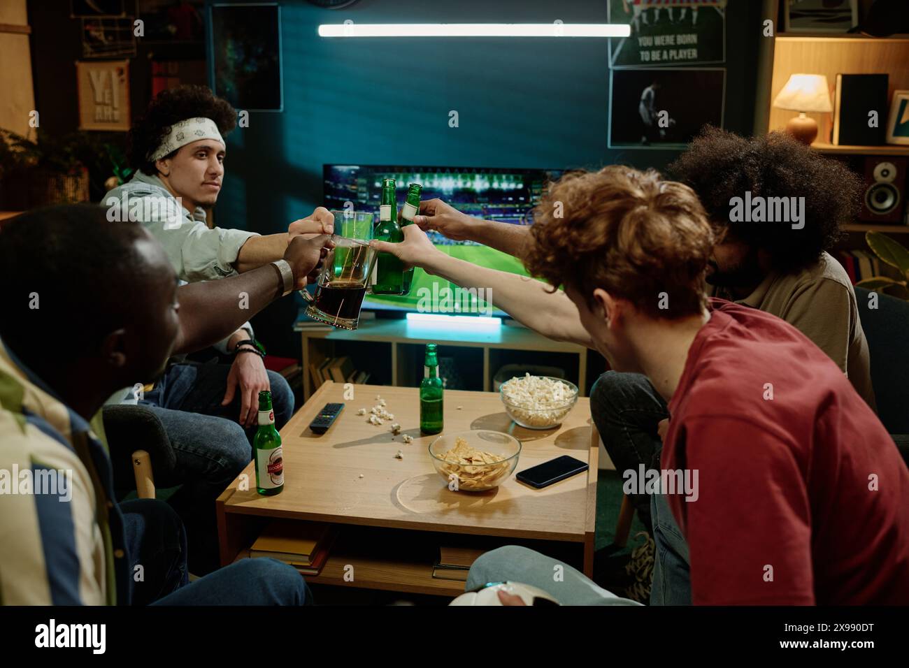 Groupe d'amis masculins ethniquement diversifiés clinquant des bouteilles avec de la bière tout en regardant le match de joueur de football à la télévision dans le salon Banque D'Images