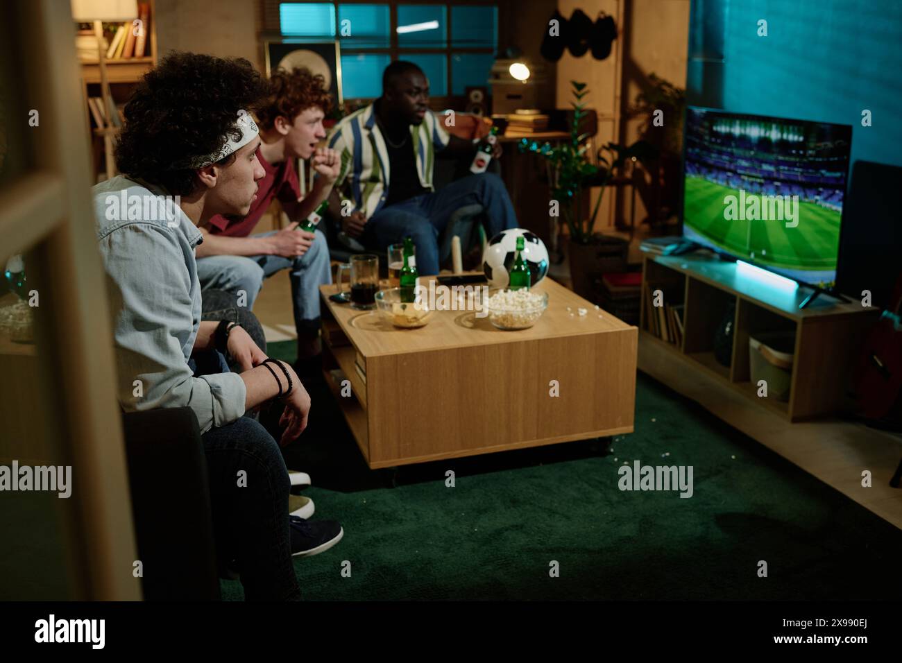 Groupe d'amis masculins multi-ethniques assis dans le salon regardant un match de football à la télévision dans la soirée, espace de copie Banque D'Images