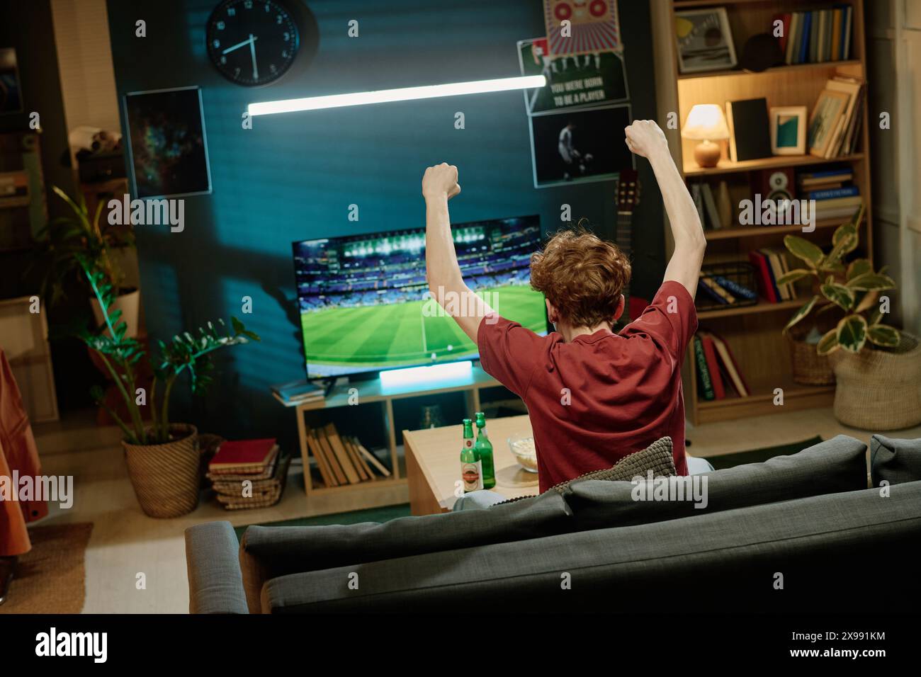 Vue arrière d'un homme aux cheveux roux méconnaissable assis avec les bras levés sur le canapé regardant un match de football à la télévision Banque D'Images