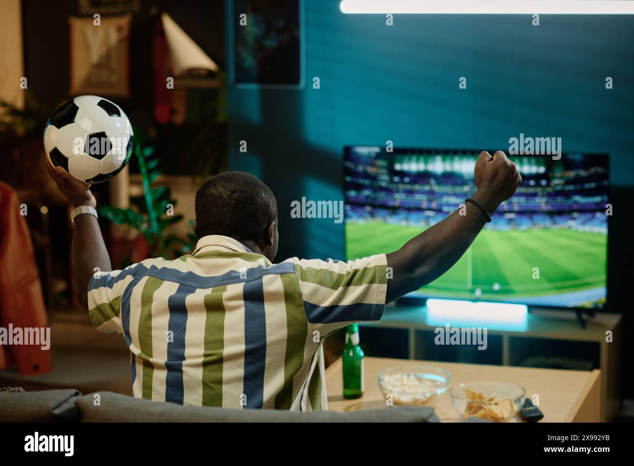 Vue arrière de fan de football afro-américain méconnaissable tenant le match de balle regardant sur la télévision dans le salon à la maison, espace de copie Banque D'Images