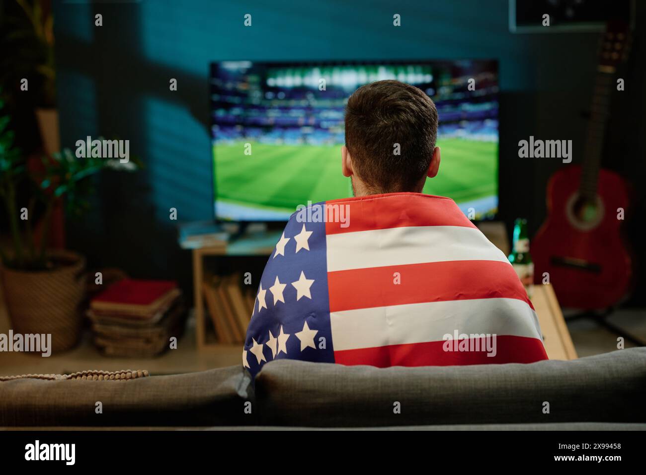 Vue arrière de l'homme méconnaissable portant drapeau assis dans le salon regardant un événement sportif à la télévision à la maison Banque D'Images