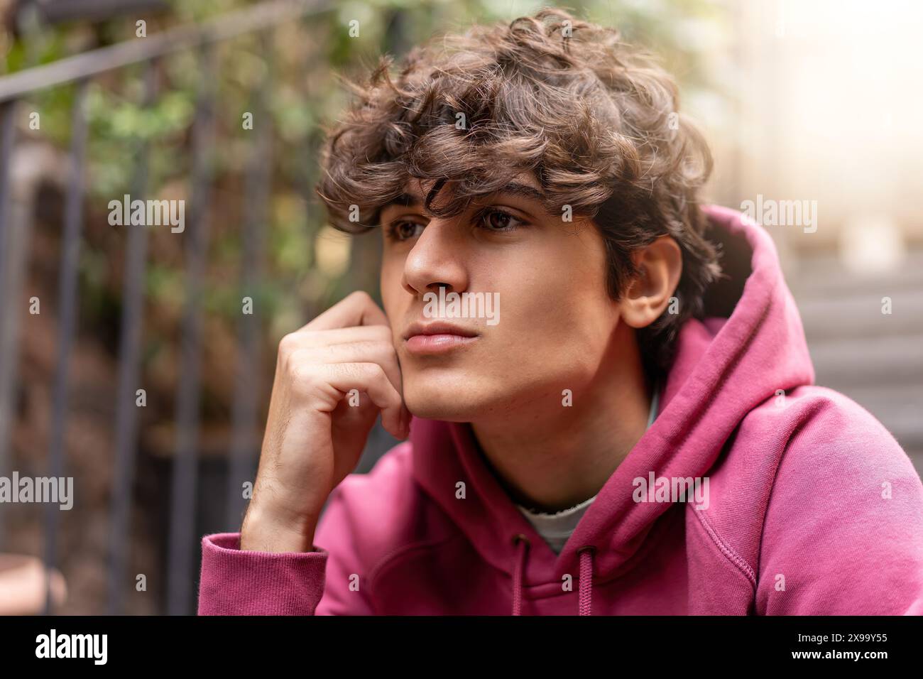 Adolescent portant un sweat à capuche rose, assis à l'extérieur et regardant réfléchi, expression pensive, mode décontractée, lumière naturelle, contemplation jeune, re Banque D'Images