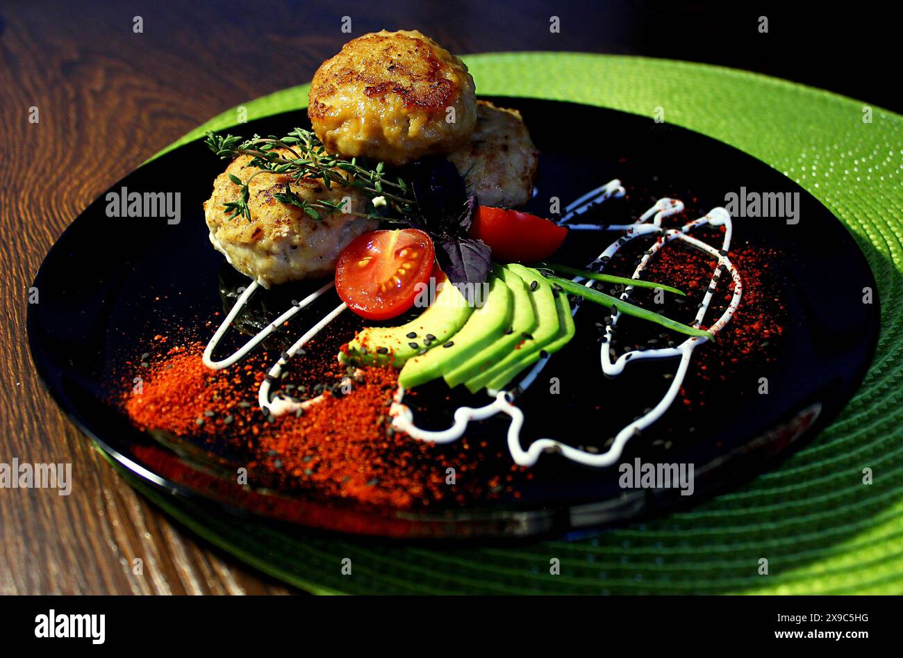 Plat gastronomique avec galettes de viande, avocat, tomate cerise et herbes confectionné sur une assiette noire avec présentation artistique Banque D'Images