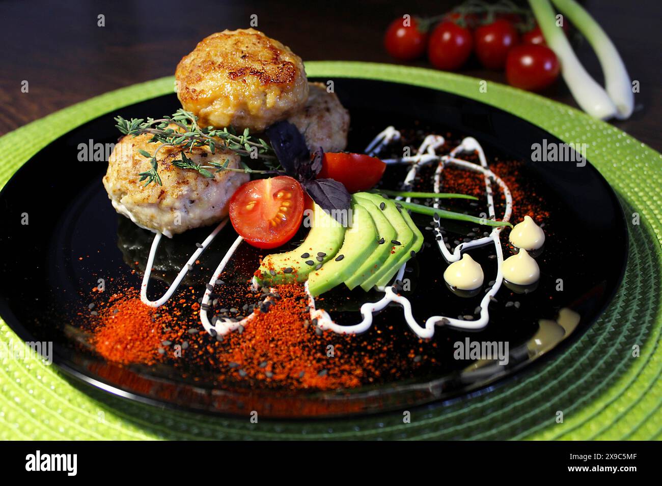 Assiette noire avec galettes de poulet, avocat, tomates cerises et sauce dans une présentation artistique avec un fond d'oignons de printemps et de tomates Banque D'Images