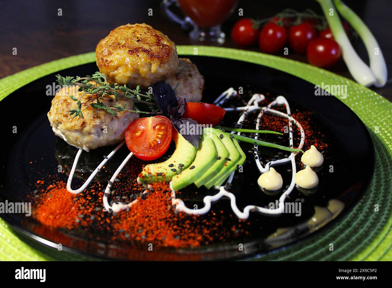 Présentation gastronomique avec des galettes de viande, des tomates, des avocats et des herbes disposées de manière créative sur une assiette noire avec des filets de sauce artistiques Banque D'Images