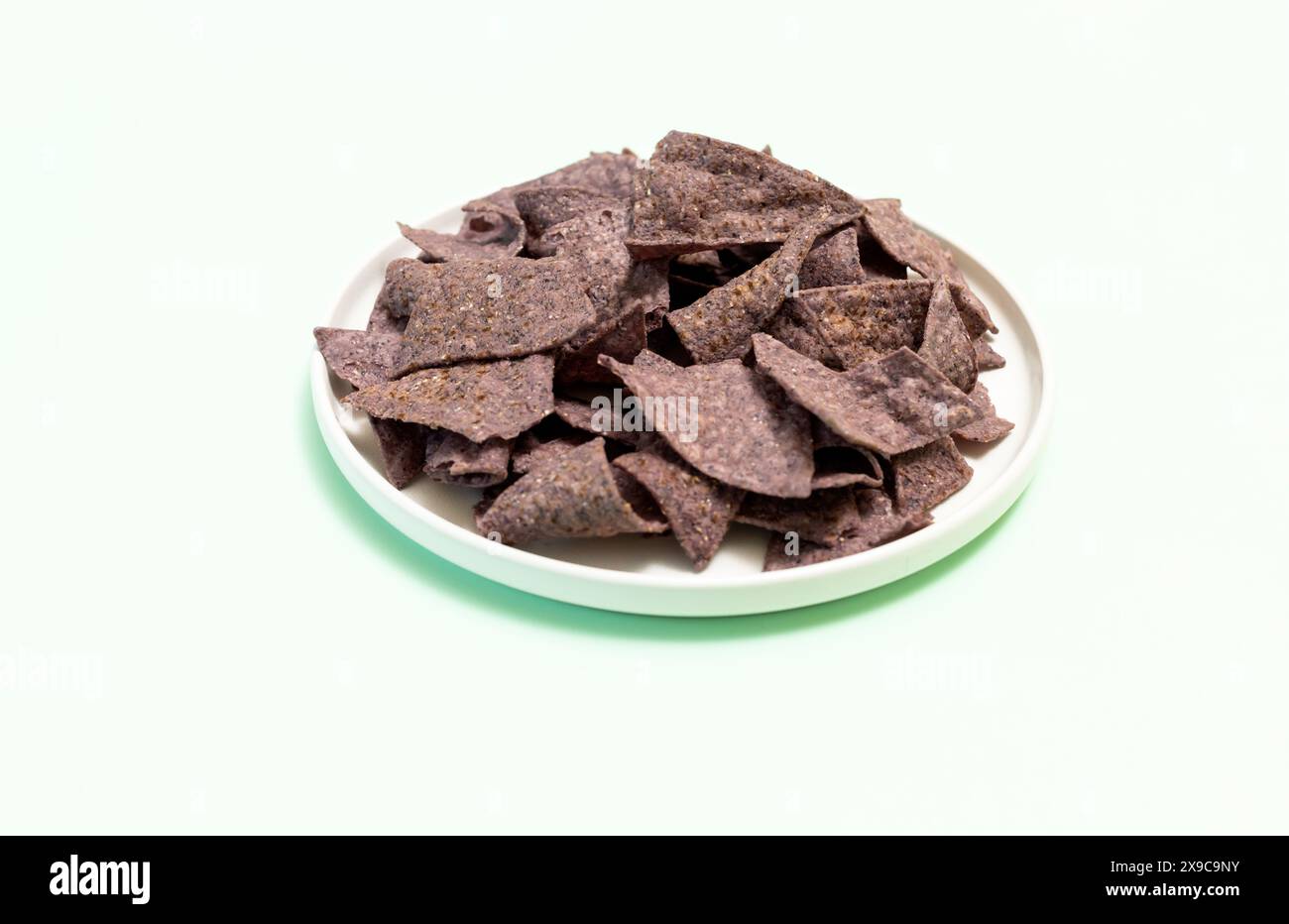 Chips de tortilla de maïs bleu organique sur assiette sur fond vert clair. Collation crunchy saine assaisonnée de sel de mer. Plan horizontal de conception. Banque D'Images