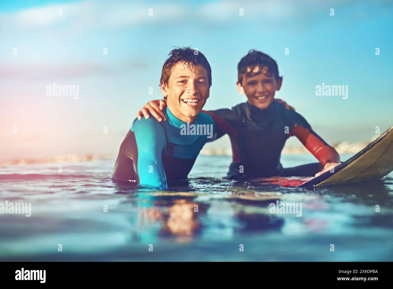 Portrait, enfants et heureux dans l'eau pour le surf, le sport et le bien-être avec planche, ciel bleu et sourire. Garçons, excités et nageant à la plage comme surfeur pour Banque D'Images