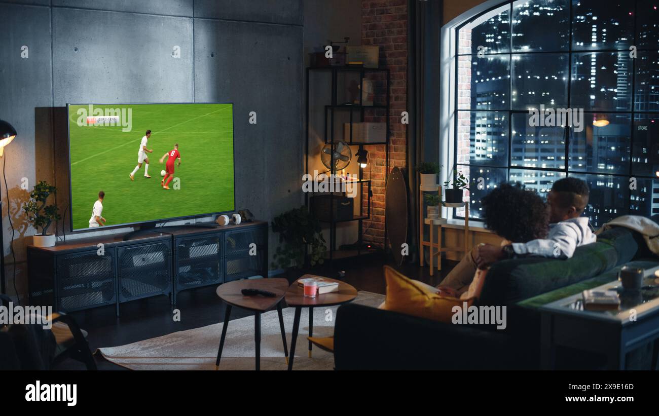 Couple de fans de football afro-américain se détendre sur un canapé, regarder un match de sport la nuit à la maison dans élégant appartement loft. Jeune homme et femme appréciant le Game of Favorite Football Club. Banque D'Images