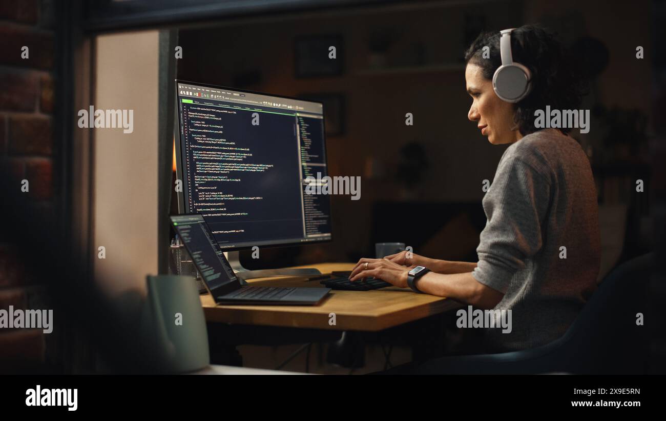 Programmeuse brésilienne travaillant sur l'ordinateur à la maison. Brillant programmateur INFORMATIQUE féminin utilisant un ordinateur de bureau tard dans la nuit. Code de programmation pour développeur professionnel. Tiré dans la fenêtre de l'extérieur. Banque D'Images