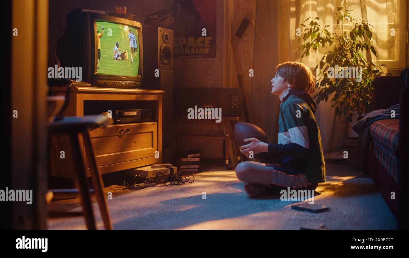 Jeune fan de sport regarde le match de football américain sur Retro TV dans sa chambre Vintage avec intérieur daté. Garçon soutenant son équipe préférée, se sentant fier lorsque les joueurs marquent un but. L'enfance nostalgique. Banque D'Images