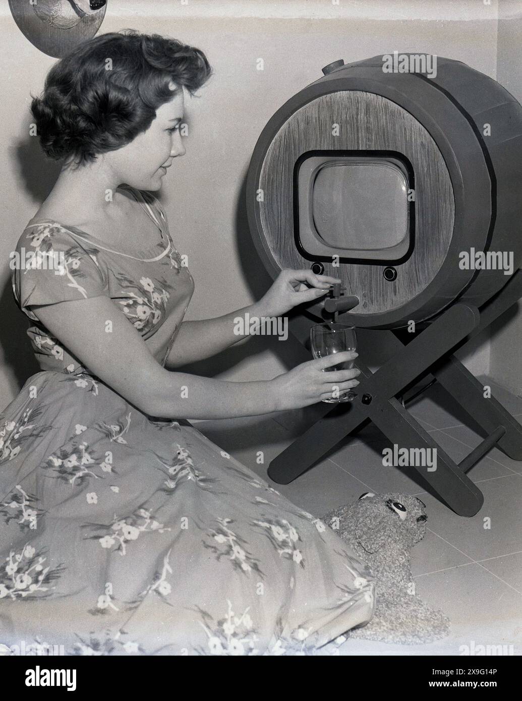 Années 1950, historique, un téléviseur en forme de tonneau avec un distributeur de boissons, une jeune dame portant une robe de l'époque, démontrant comment cela fonctionne, tenant un verre sous l'écran, versant une boisson, USA. Banque D'Images Années 1950, historique, un téléviseur en forme de tonneau avec un distributeur de boissons, une jeune dame portant une robe de l'époque, démontrant comment cela fonctionne, tenant un verre sous l'écran, versant une boisson, USA. Banque D'Images