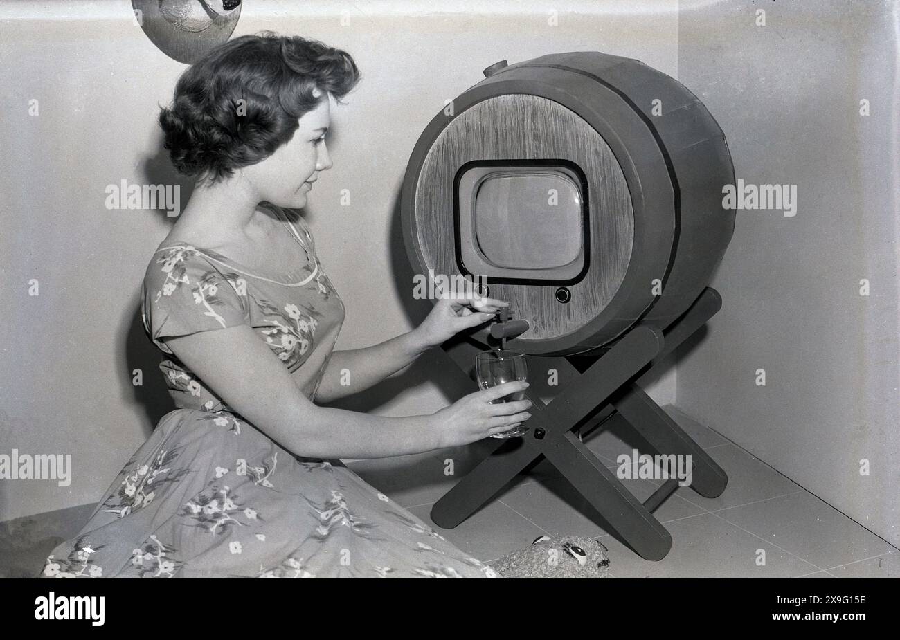 Années 1950, historique, un téléviseur en forme de tonneau avec un distributeur de boissons, une jeune dame portant une robe de l'époque, démontrant comment cela fonctionne, tenant un verre sous l'écran, versant une boisson, USA. Banque D'Images Années 1950, historique, un téléviseur en forme de tonneau avec un distributeur de boissons, une jeune dame portant une robe de l'époque, démontrant comment cela fonctionne, tenant un verre sous l'écran, versant une boisson, USA. Banque D'Images