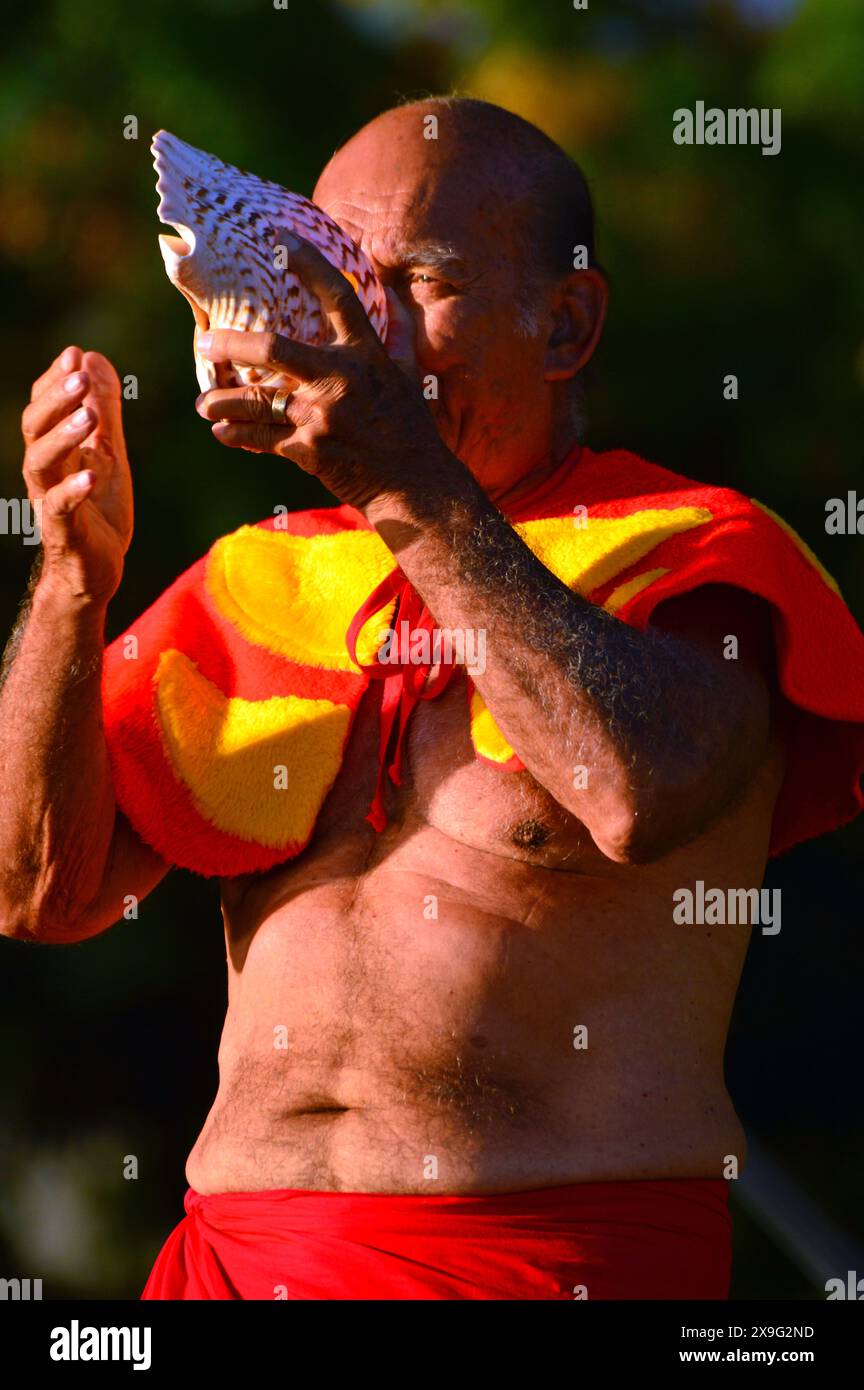 Un homme âgé, vêtu d'une tenue hawaïenne traditionnelle signale le coucher du soleil en soufflant dans une coquille de conque sur la plage de Waikiki à Hawaï Banque D'Images