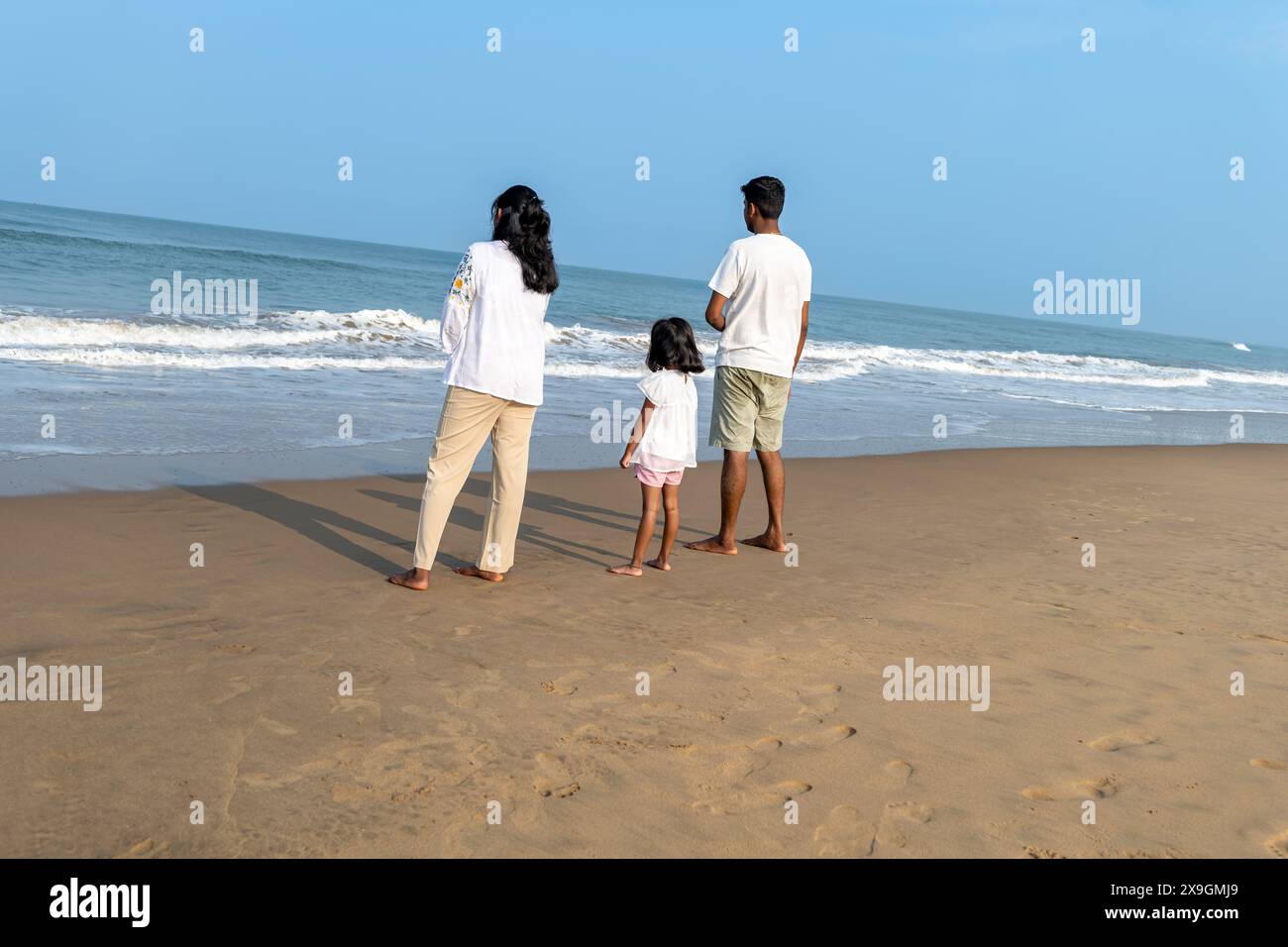 Une famille joyeuse – père, mère et fille – embrassant la plage ensoleillée, leurs rires portés par les douces vagues. Banque D'Images