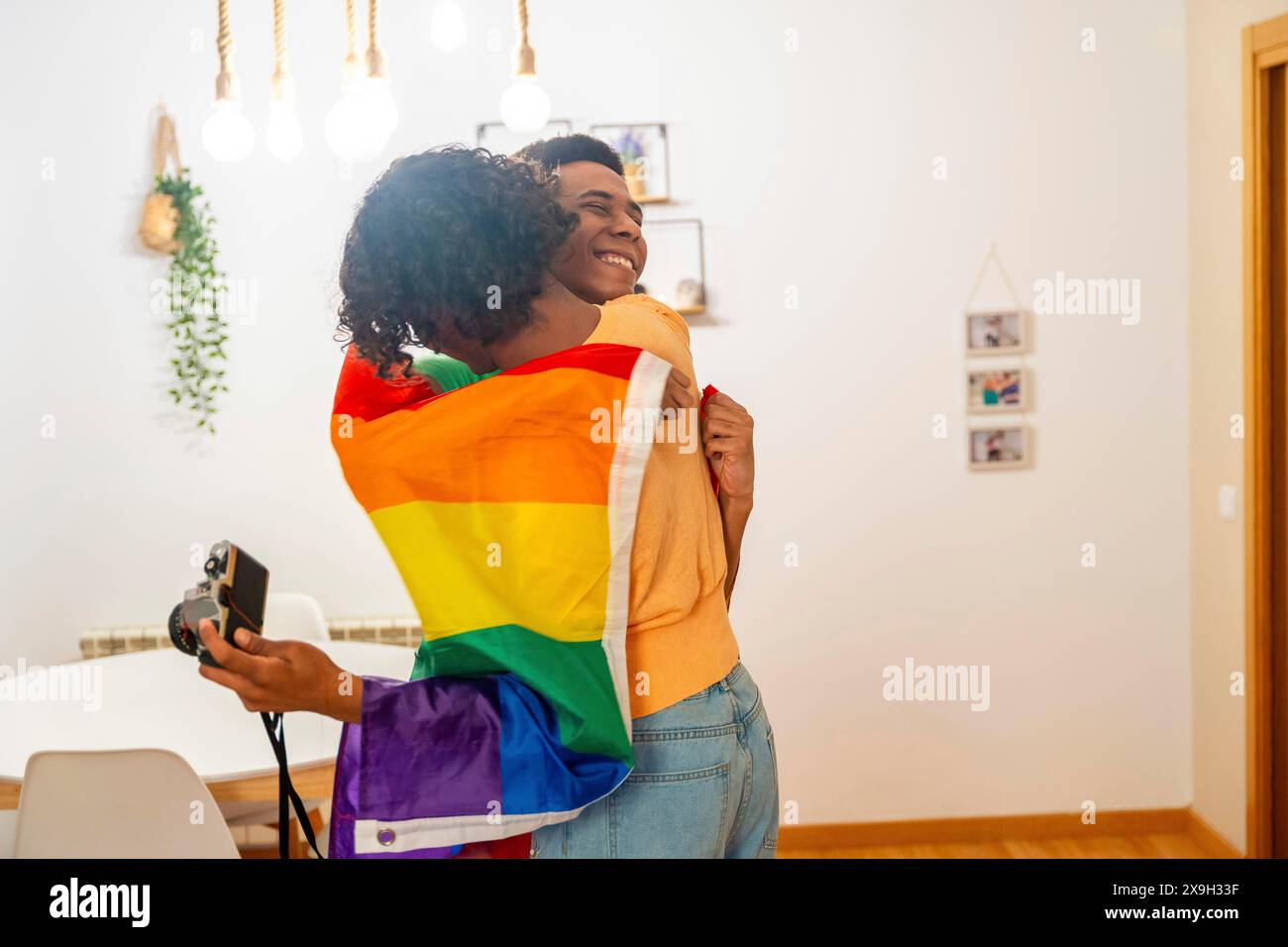 Jeune couple gay latin enveloppé dans le drapeau LGBT tout en prenant des photos dans l'appartement Banque D'Images