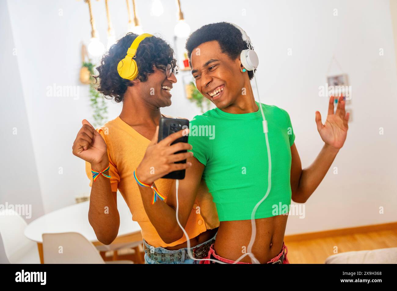 Jeune couple masculin gay latin dansant en écoutant de la musique avec des écouteurs et un mobile debout à la maison Banque D'Images