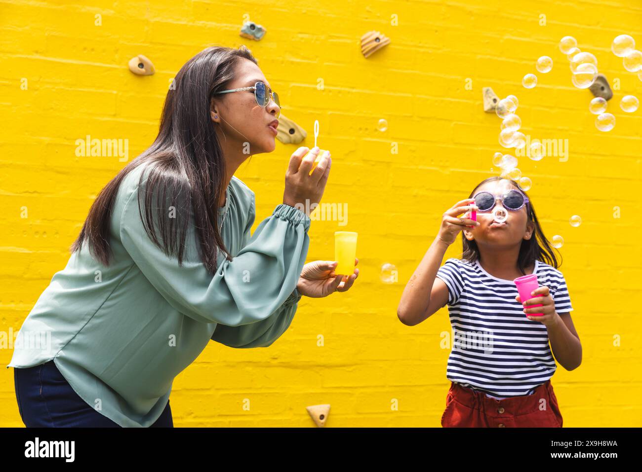 Une femme et une jeune fille biraciale soufflent des bulles dehors contre un mur jaune Banque D'Images