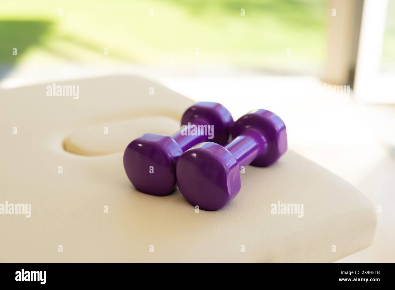 À la maison, deux haltères violets reposant sur une surface rembourrée blanche dans une pièce lumineuse. Lumière du soleil qui traverse une fenêtre, projette des ombres douces et illuminati Banque D'Images