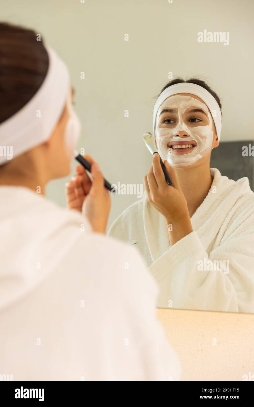 À la maison, jeune femme caucasienne appliquant un masque facial tout en se regardant dans le miroir. Dans une salle de bain confortable, profiter d'une journée spa relaxante avec un éclairage doux et de la suie Banque D'Images