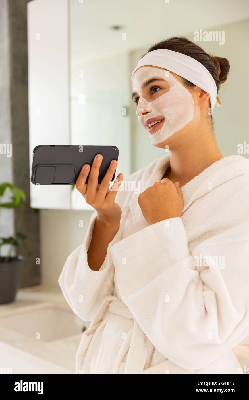 Femme caucasienne à la maison portant un masque facial, tenant un téléphone dans une salle de bain semblable à un spa. Elle porte un peignoir blanc, profitant d'un moment de détente et de détente Banque D'Images