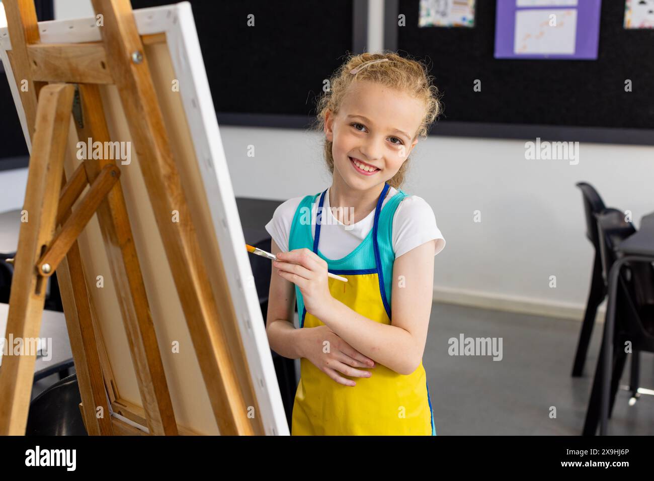 Fille caucasienne avec des peintures de cheveux blonds sur un chevalet à l'école, portant un tablier jaune Banque D'Images