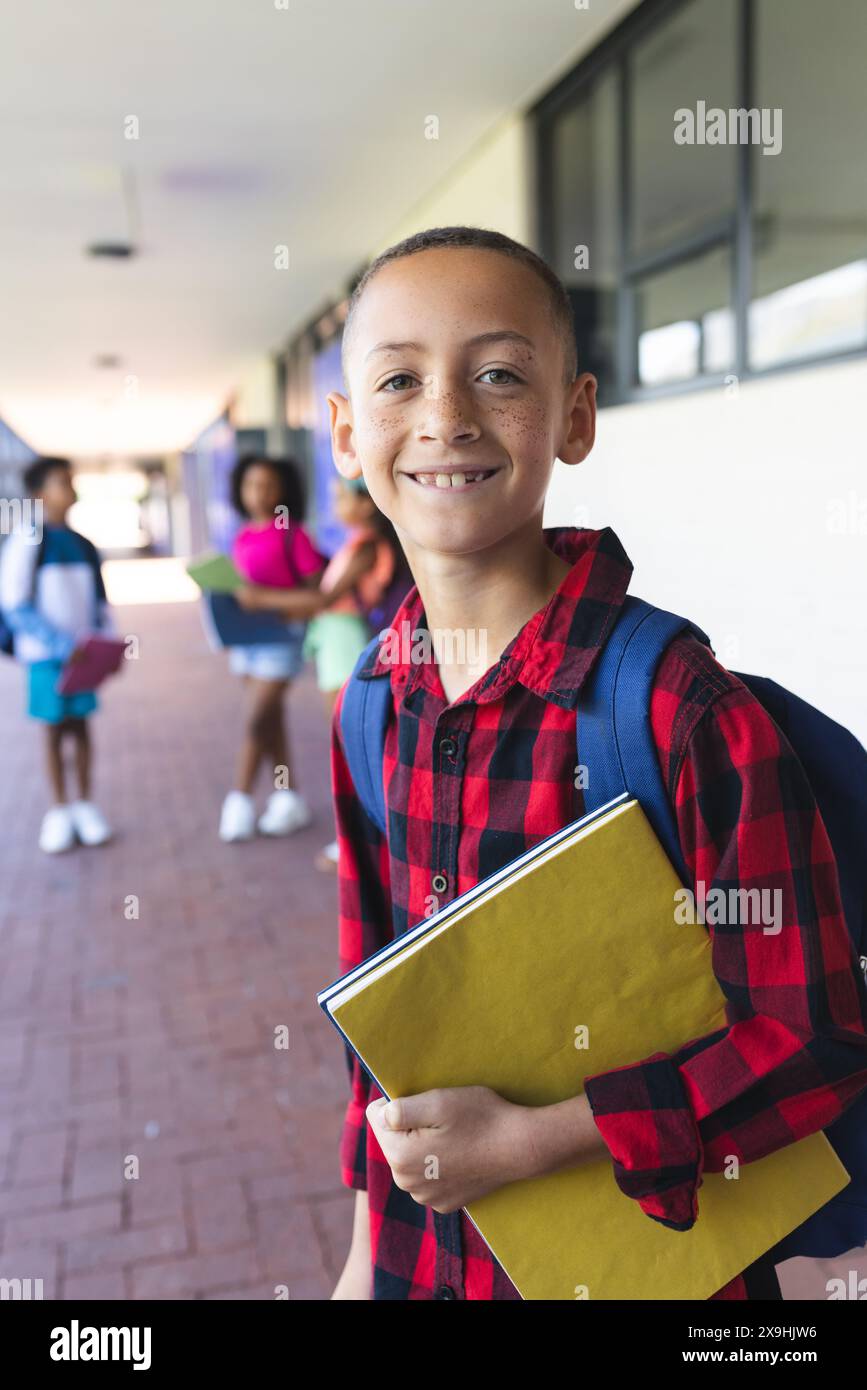 Garçon biracial avec un sac à dos et un carnet jaune sourit à l'école Banque D'Images
