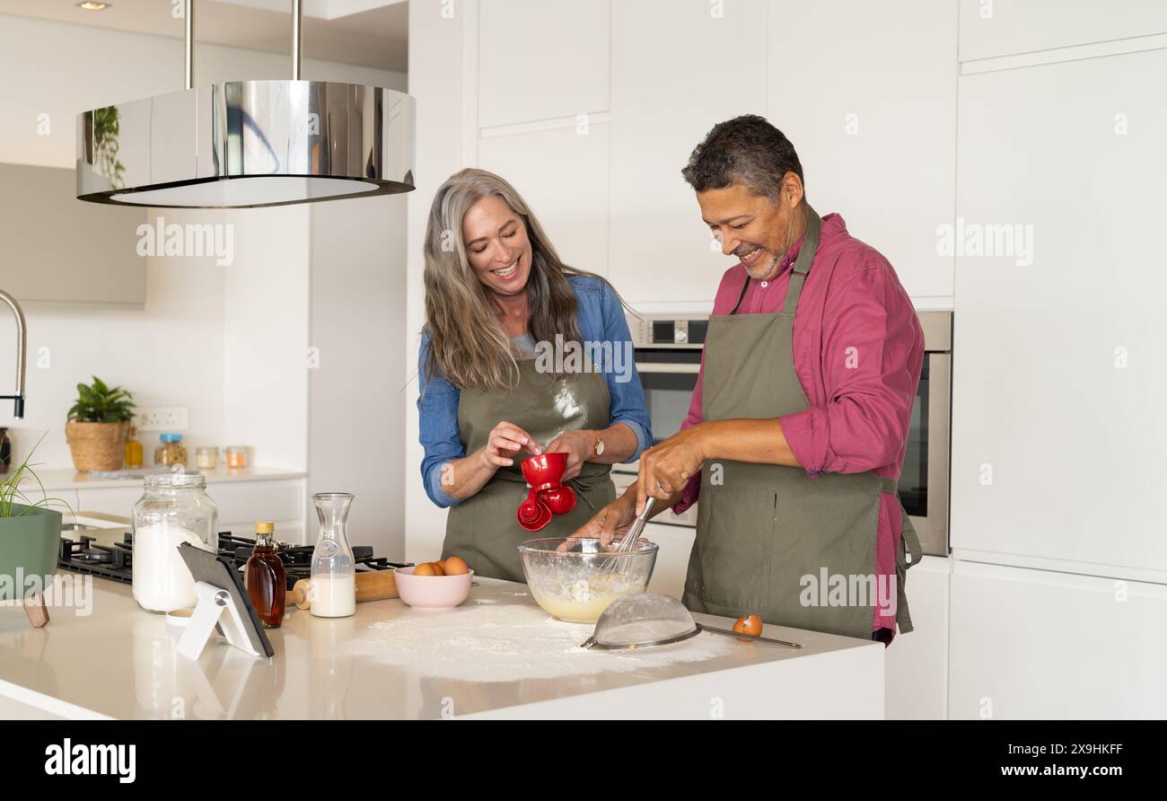 À la maison, couple senior diversifié portant des tabliers cuisant ensemble dans une cuisine moderne. Femme caucasienne avec de longs cheveux gris et homme biracial avec un court foncé Banque D'Images