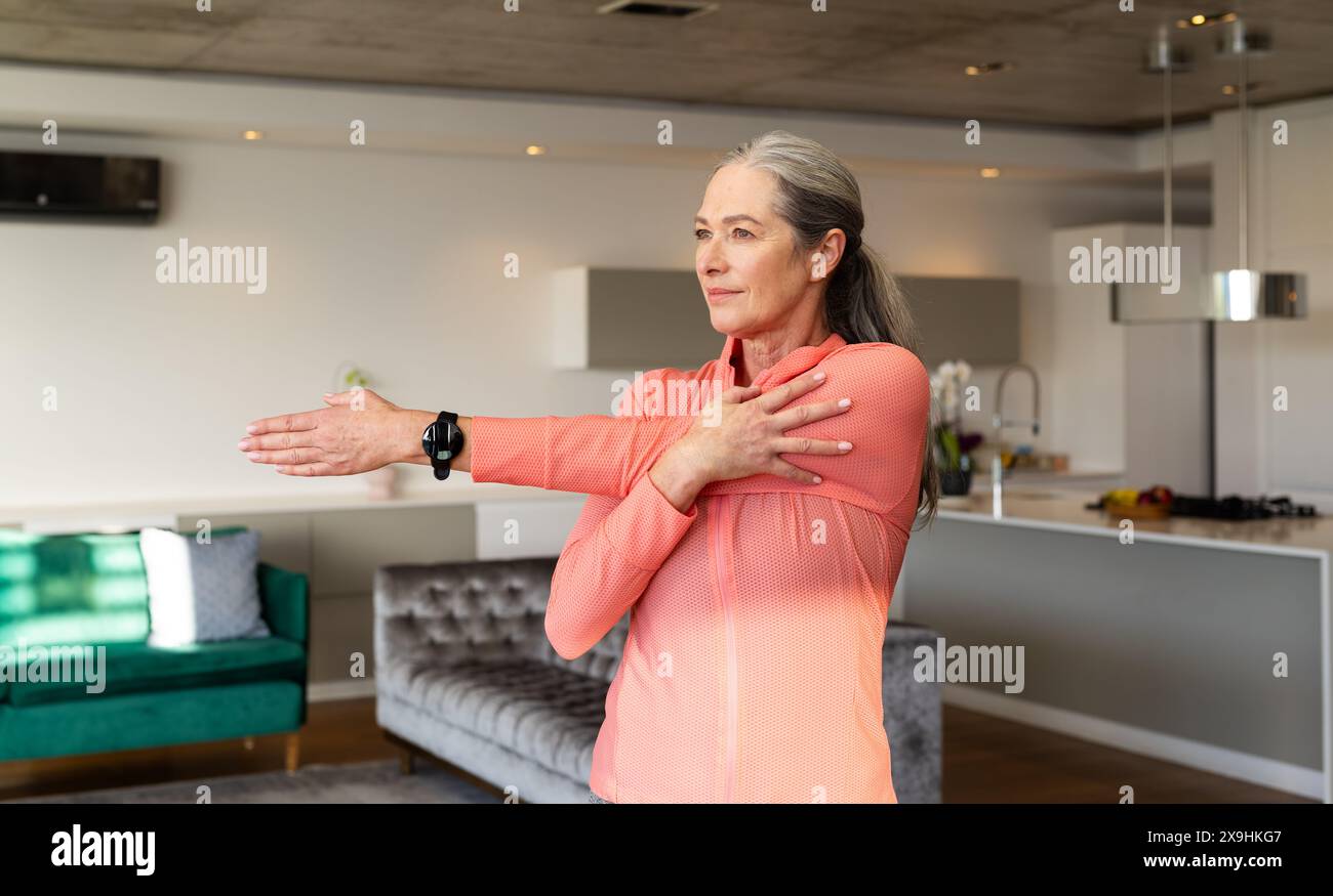 À la maison, une femme âgée caucasienne aux longs cheveux gris s'étire dans un salon moderne. Portant un haut rose à manches longues et une smartwatch noire, look Focu Banque D'Images