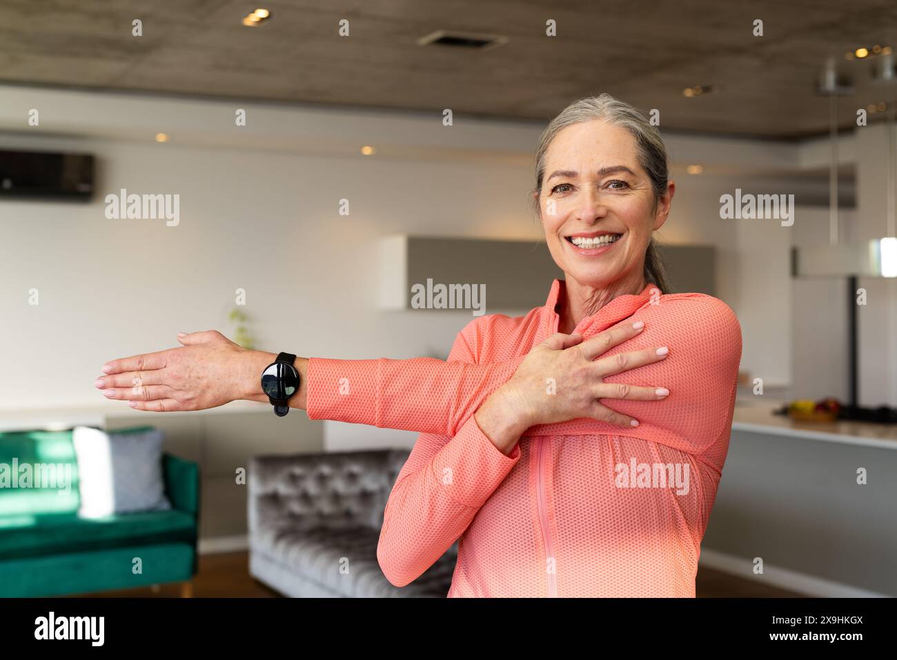 À la maison, femme âgée caucasienne avec les cheveux gris et les yeux bleus s'étirant. Portant un haut de sport rose à manches longues et une montre de fitness noire, souriant w Banque D'Images