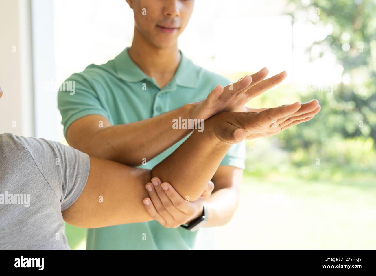 Un jeune physiothérapeute masculin biracial aide une femme biraciale senior à faire des exercices à la maison. Ils sont dans une pièce lumineuse et ensoleillée avec une vue sur un g luxuriant Banque D'Images