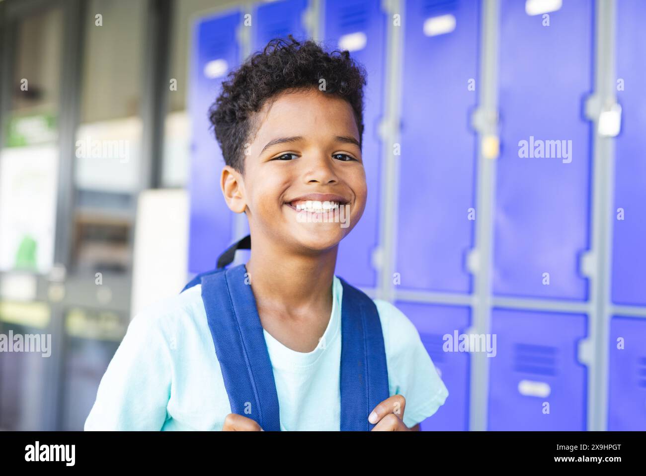 Garçon biracial aux cheveux bouclés sourit devant des casiers bleus à l'école, portant un sac à dos Banque D'Images